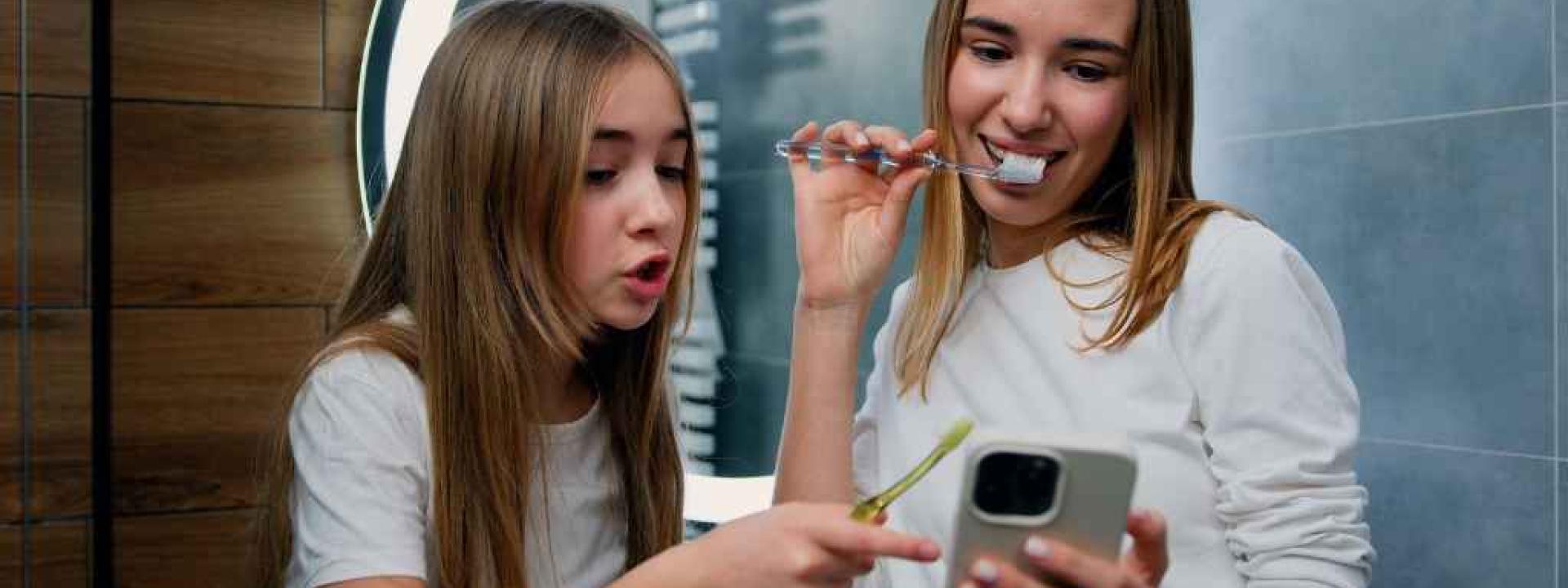 Two young girls smiling, brushing their teeth and looking at a smartphone